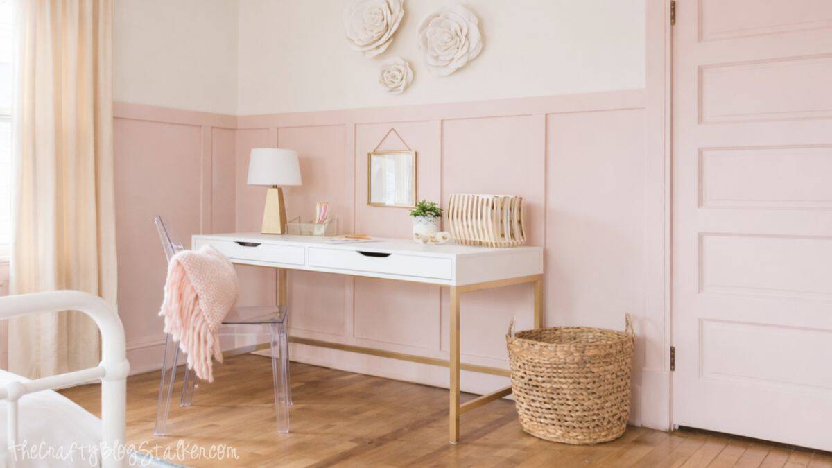 Styled girls bedroom desk area with a white desk, clear chair, decorative wall flowers, small mirror, table lamp, file organizer, and woven floor basket.