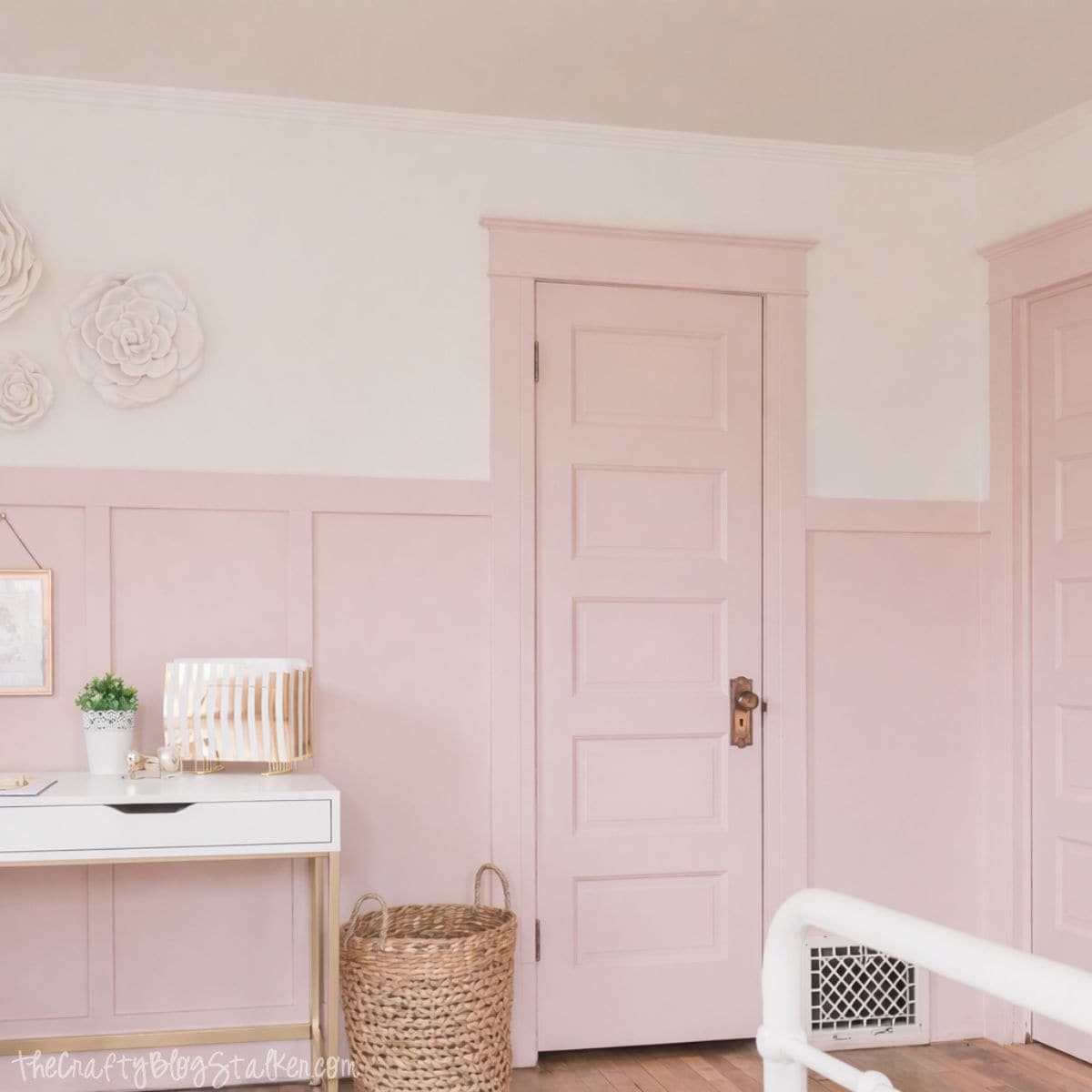 Girls bedroom wall with paneled wainscoting, a door, woven basket, and a white desk styled with decor and wall art.