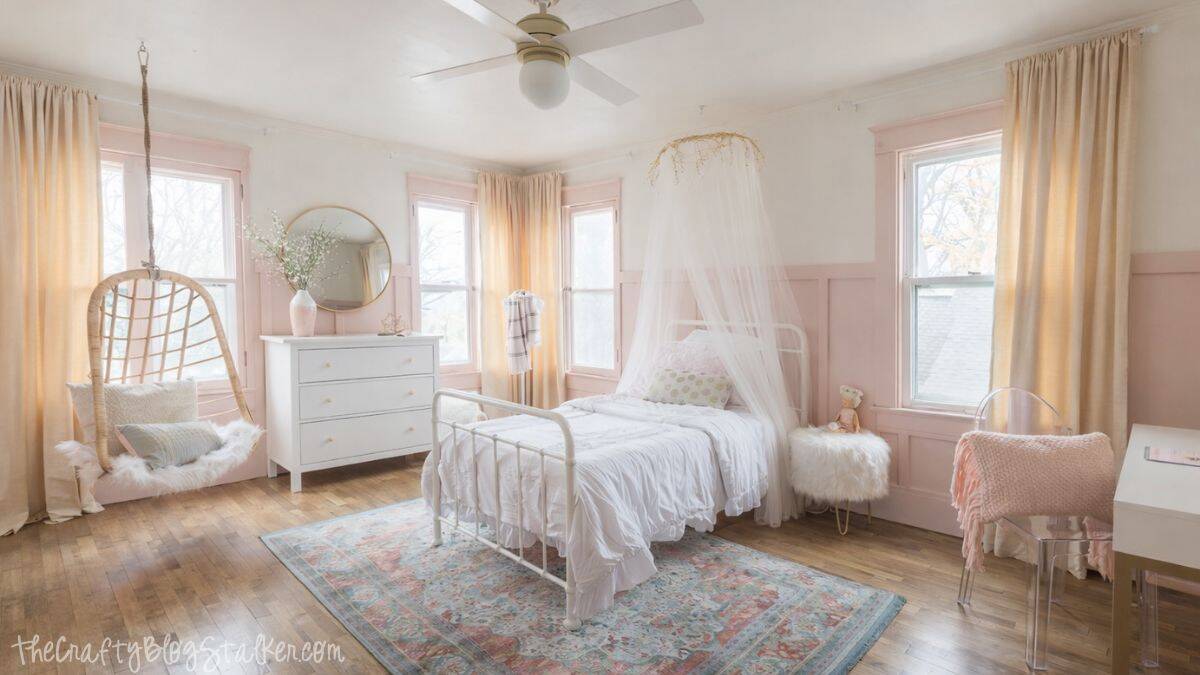 Wide view of a girls bedroom with a white metal bed and canopy, hanging wicker chair, dresser, desk, soft rug, and large windows letting in natural light.
