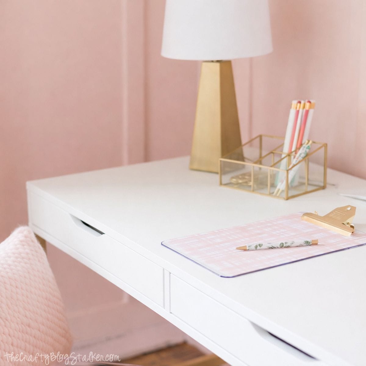 Close-up of a modern desk with a table lamp, gold pencil holder, clipboard, and notepad, styled as a girls bedroom workspace with paneled walls.