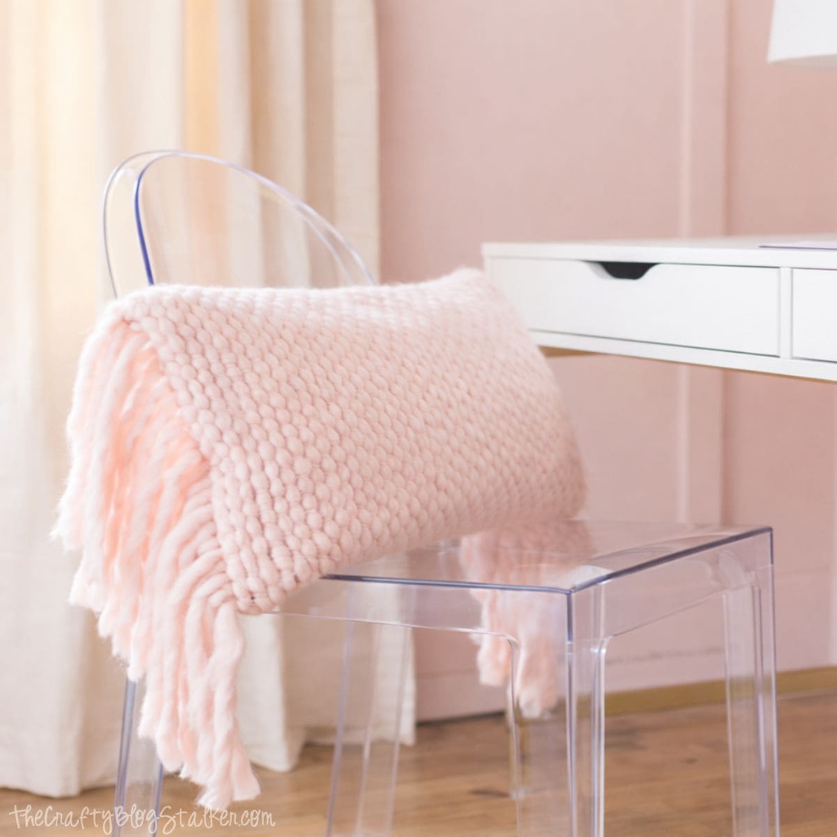 Clear acrylic chair with a soft knitted throw draped over the back, tucked under a white desk in a girls bedroom with light wood floors and soft curtains.