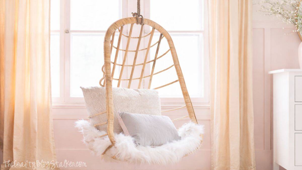 Hanging wicker chair with a soft faux fur seat and layered pillows, suspended in front of a window with curtains in a cozy girls bedroom.