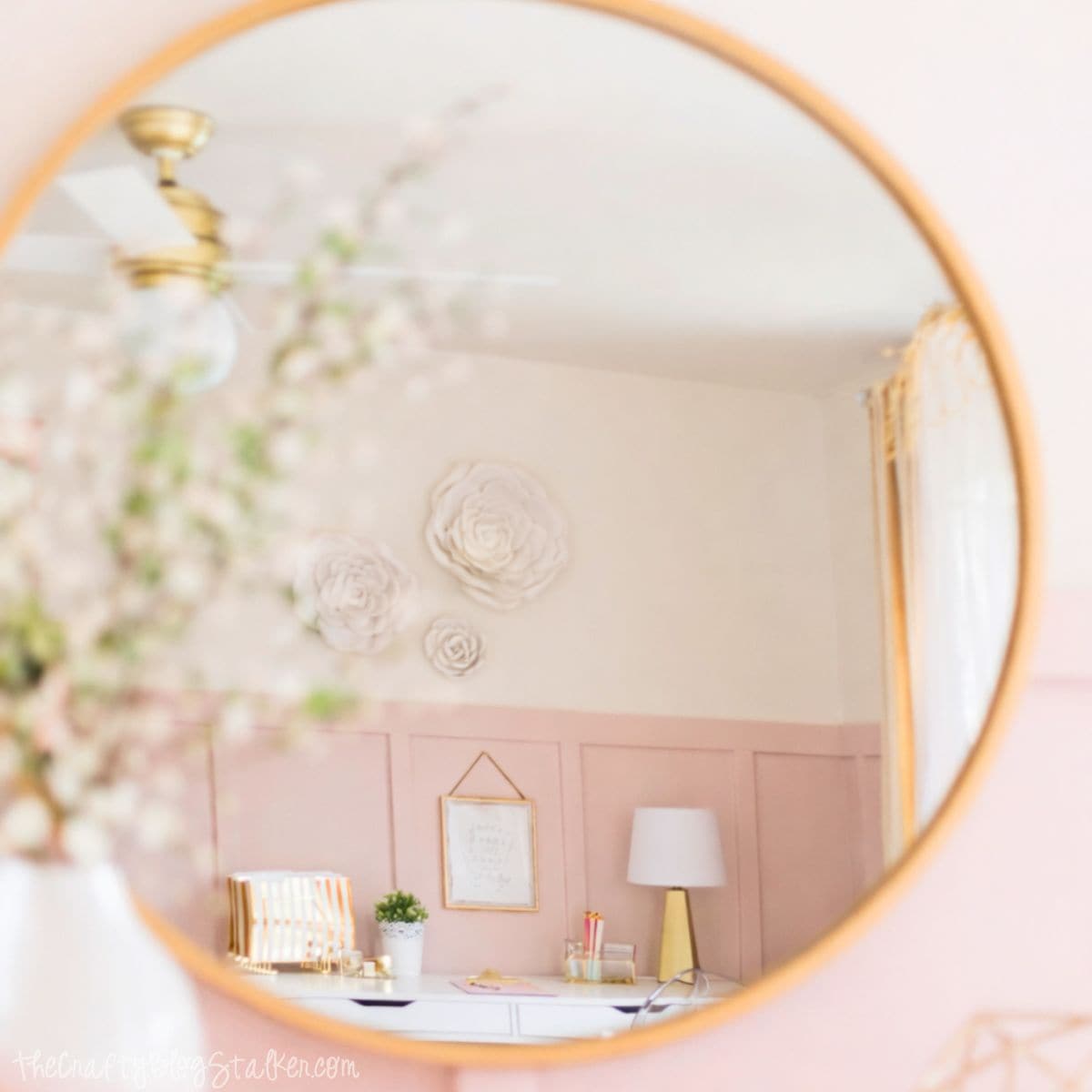 Round wall mirror reflecting a girl's bedroom desk with a lamp, wall art, floral decor, and paneled walls, creating a soft, styled bedroom vignette.
