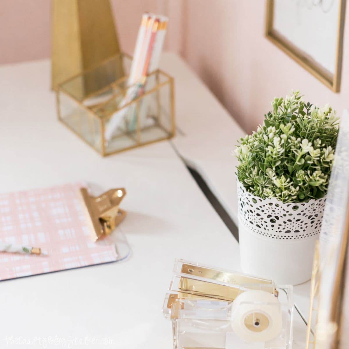 Styled desk surface with a small potted plant, clear tape dispenser, gold stationery holder, clipboard, and pencils arranged on a white desk in a girls bedroom.