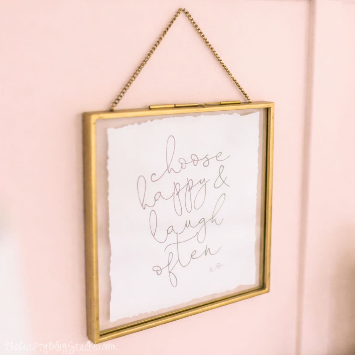 Gold-framed wall art hanging on a paneled bedroom wall, featuring a handwritten quote displayed in a minimalist frame with a chain hanger.