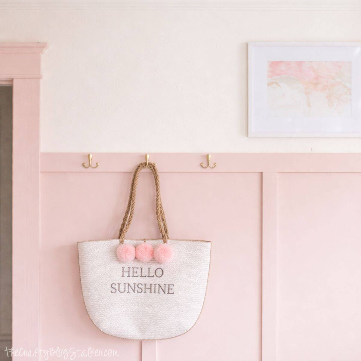 Woven tote bag with rope handles and pom-pom accents hanging on wall hooks above paneled wainscoting, displayed as decorative storage in a bedroom.