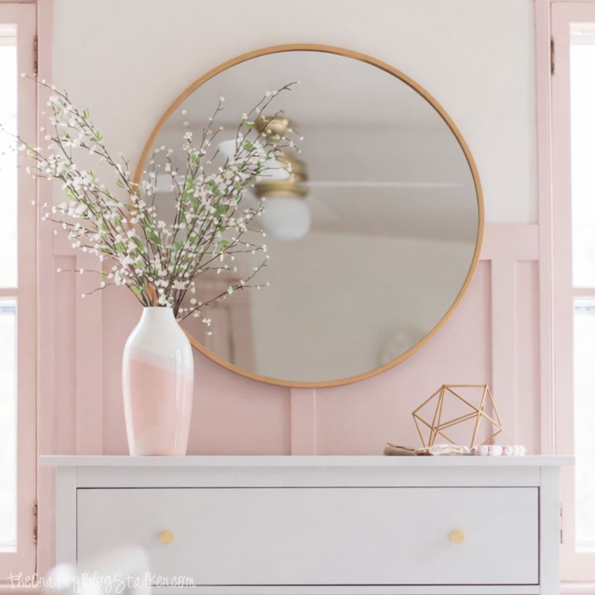 White dresser topped with a vase of branches and decor, styled beneath a round wall mirror in a soft, feminine bedroom setting.