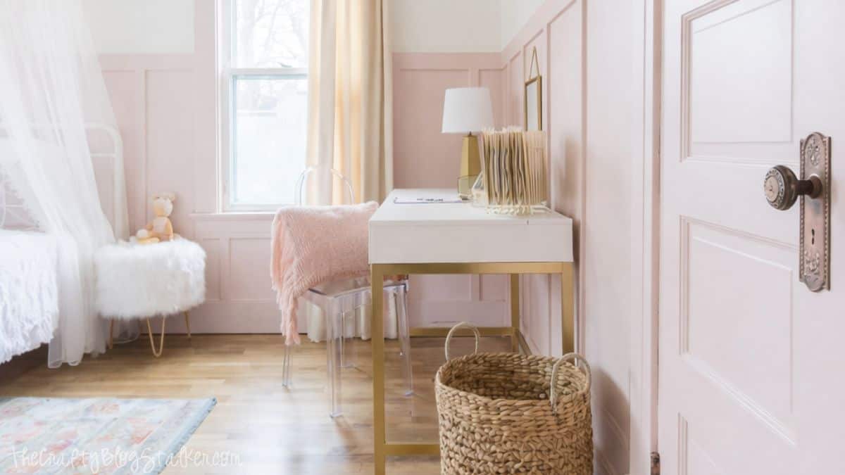 Girl's bedroom with a small desk and chair, bed with canopy, window with curtains, table lamp, woven basket, and soft decor arranged along paneled walls.