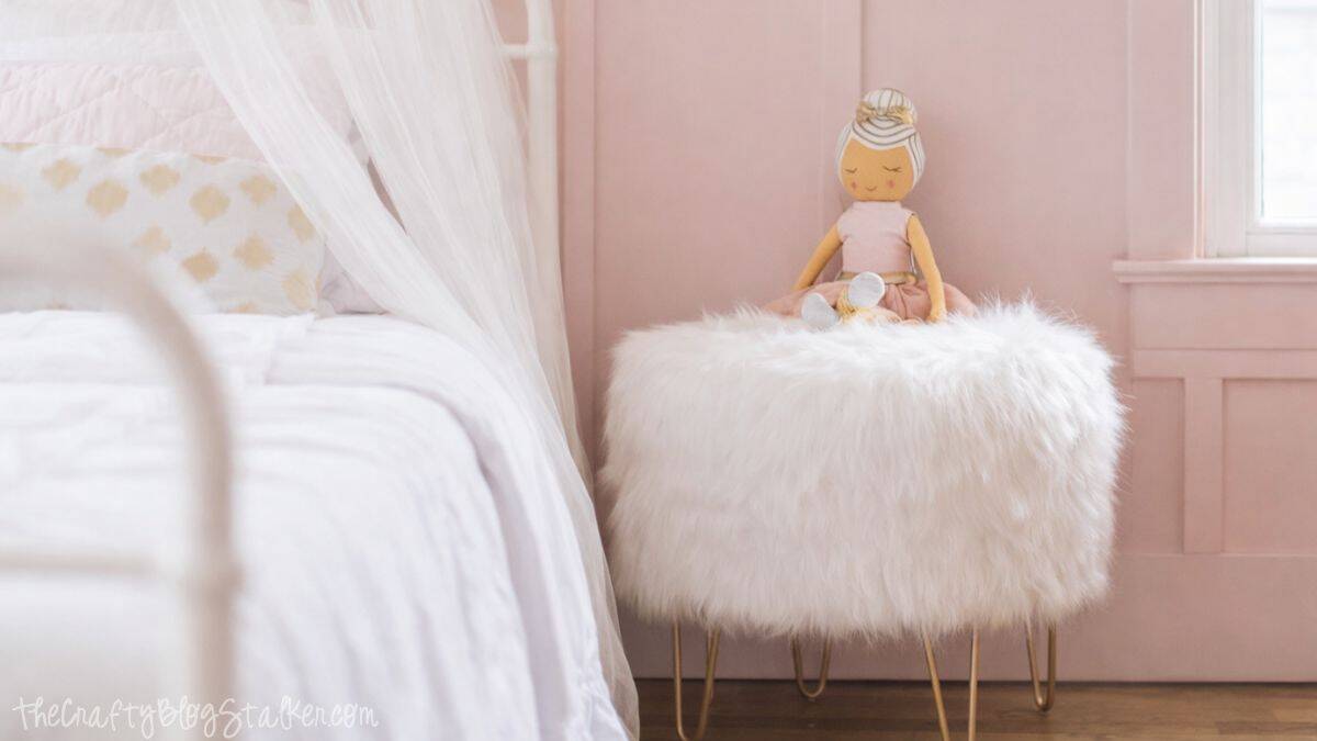 Stuffed doll sitting on a fluffy upholstered stool beside a white metal bed with sheer canopy curtains in a softly styled girls bedroom.