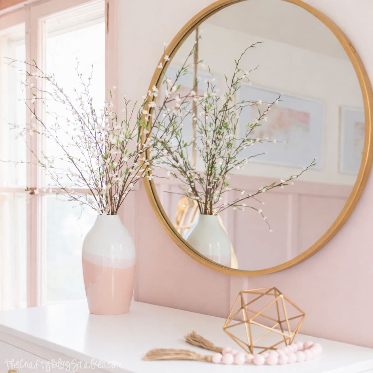 Round wall mirror above a white dresser, styled with a ceramic vase holding faux branches, a geometric decor object, and a beaded garland in a softly decorated bedroom.
