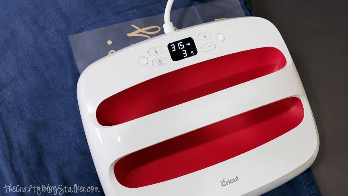 Cricut EasyPress set to 315°F applying heat to a metallic iron-on design on a blue fabric, following the recommended heat transfer settings for polyester.