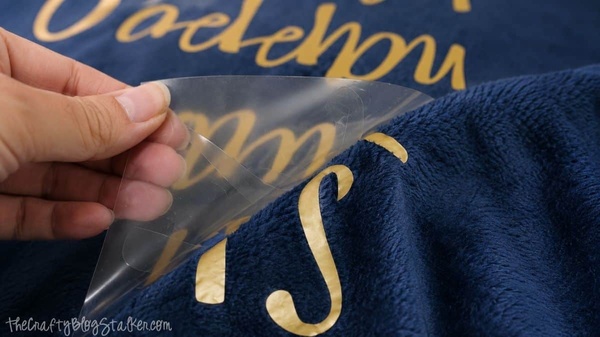 Peeling the clear transfer liner from a gold iron-on design applied to a soft blue blanket, revealing the finished text on the plush fabric.