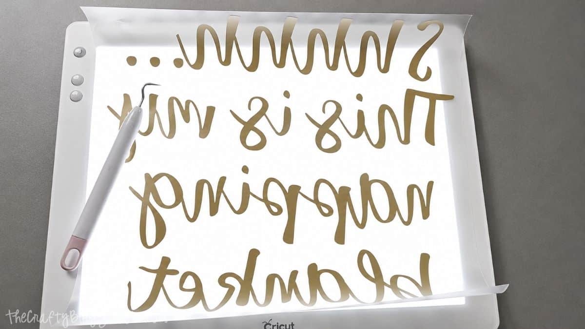 Gold iron-on vinyl with reversed text "Shhhh... This is my napping blanket" placed on a Cricut BrightPad, with a weeding tool beside it for design preparation.