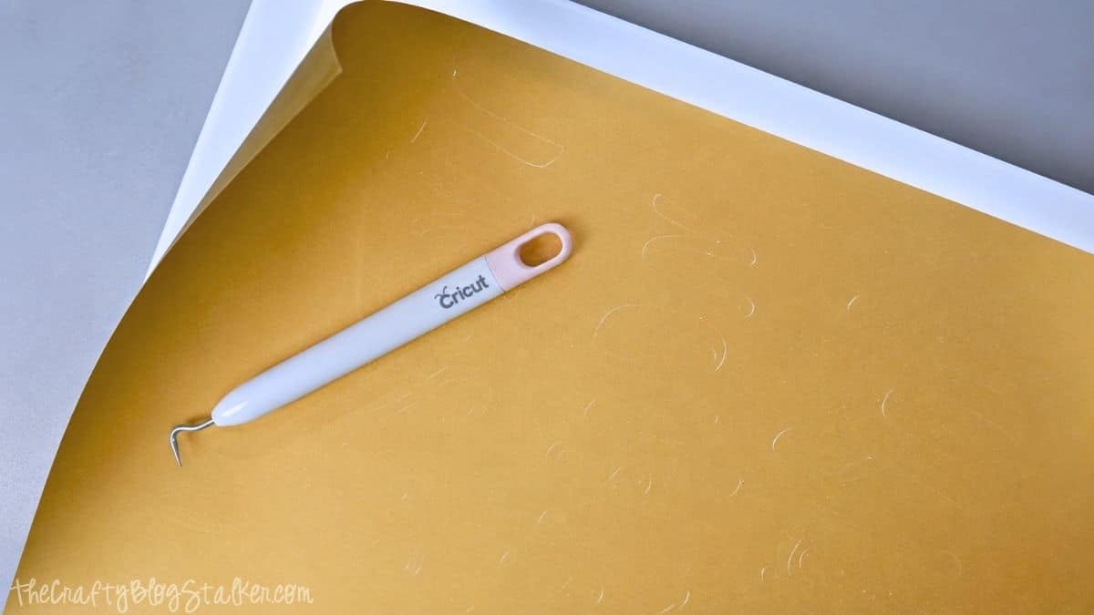 Cricut weeding tool resting on a sheet of gold heat transfer vinyl with excess vinyl partially removed for an HTV blanket project.