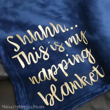 A plush blue blanket with gold lettering that reads, "Shhhh... This is my napping blanket," displayed on a dark surface.