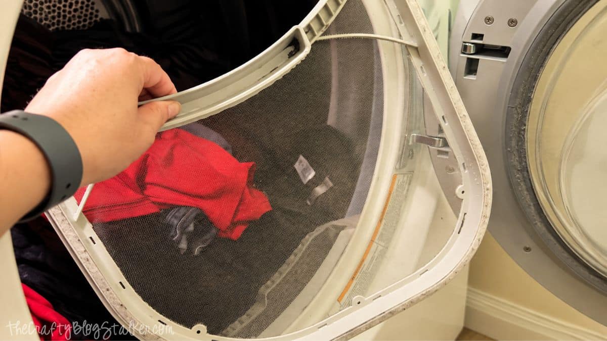 Hand holding a clean dryer lint screen in front of an open clothes dryer filled with laundry.