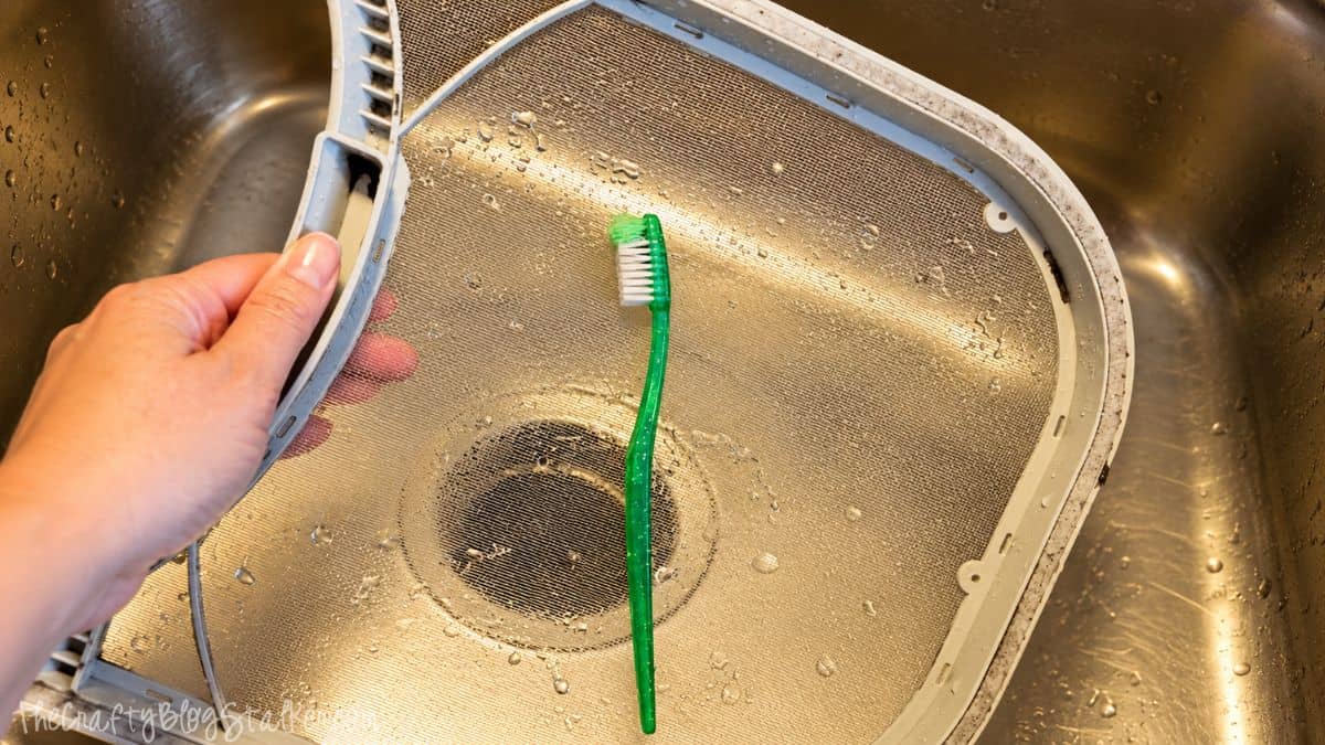 Clean dryer lint screen in a stainless steel sink with a green toothbrush resting on the mesh after scrubbing away built-up residue.
