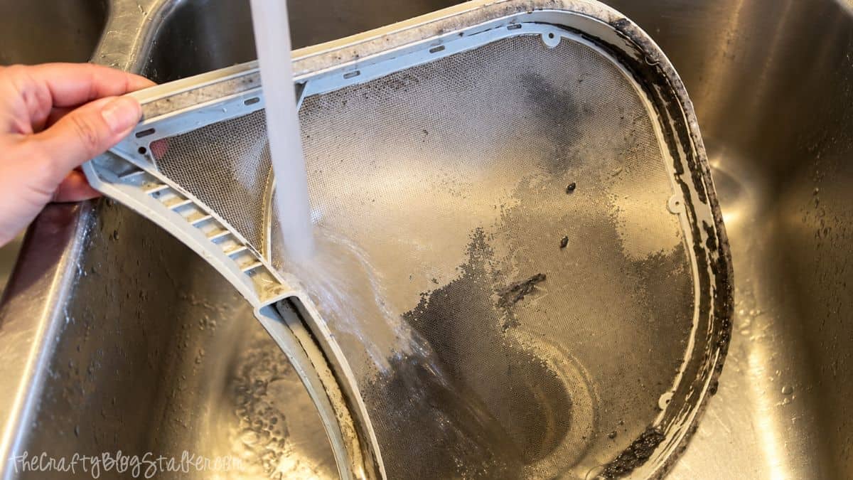 Dryer lint screen being rinsed under running water in a stainless steel sink to remove lint, dust, and built-up residue.