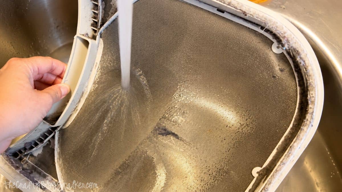 Dryer lint screen being rinsed under running water in a kitchen sink to remove built-up lint and residue.