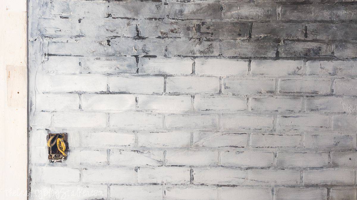 Dry brushed faux brick wall with a layered, weathered finish and visible mortar lines, shown mid project with an exposed electrical box.