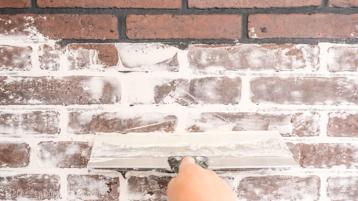 Applying joint compound to a faux brick wall using a wide putty knife to create a distressed, painted brick texture.