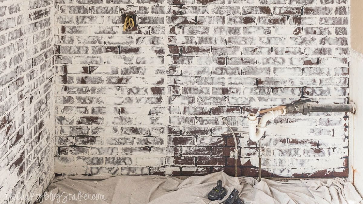 Faux brick wall coated with joint compound to create a whitewashed brick look, showing exposed plumbing and drop cloth on the floor during the DIY process.