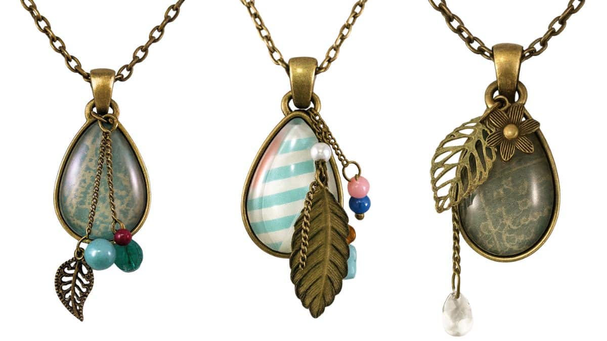 Three handmade teardrop pendant necklaces with glass cabochons, bronze chains, and charms like beads, leaves, and flowers in different designs.