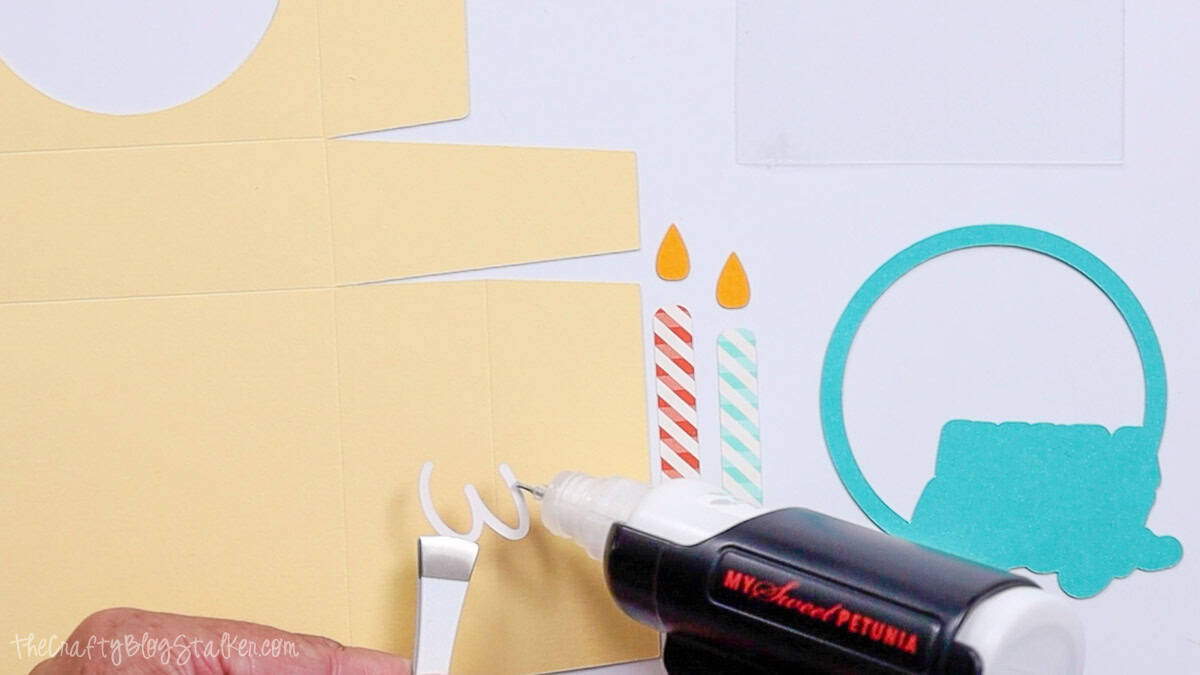 A hand uses a glue pen to apply adhesive to a small white paper cutout, with other colorful paper pieces for a gift box project arranged nearby.