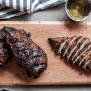 Grilled balsamic chicken breasts on a wooden cutting board, one whole and one sliced, with a metal cup of dressing and striped cloth in the background.