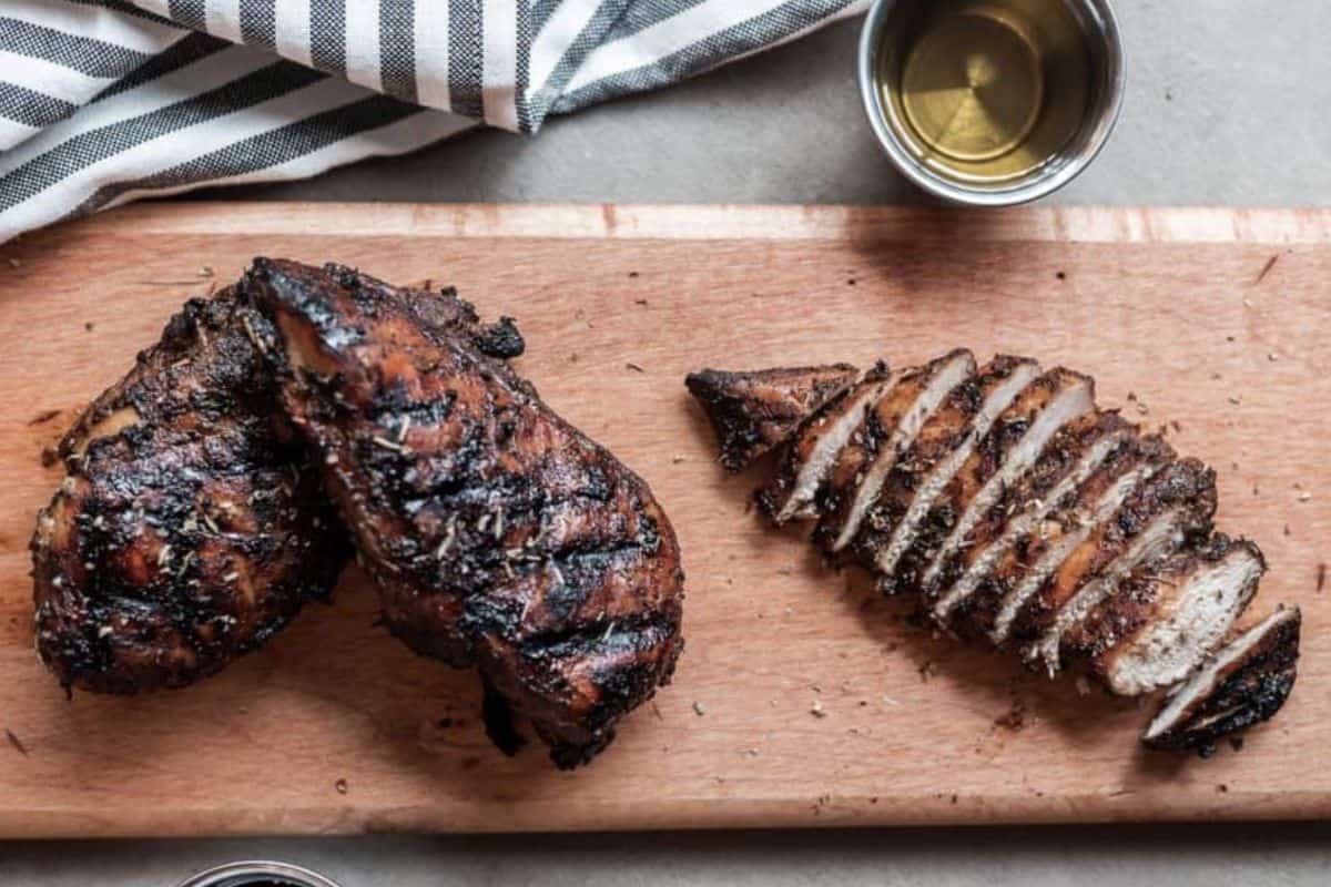 Grilled balsamic chicken breasts on a wooden cutting board, one whole and one sliced, with a metal cup of dressing and striped cloth in the background.