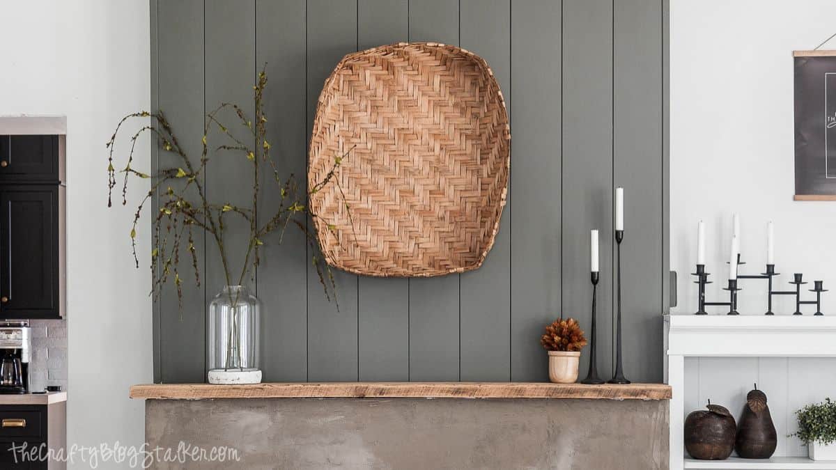 Modern farmhouse mantel decorated with a large woven basket, glass vase with branches, black candlesticks, and a small fall arrangement.