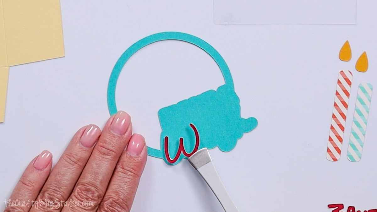 A hand holds a teal paper circle piece while using tweezers to place a red “W” cutout onto it, with other paper candle pieces nearby.