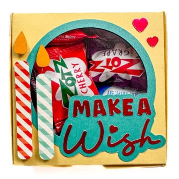 Yellow gift box with a clear window, filled with Zotz candy, decorated with striped candles, hearts, and the phrase “Make a Wish” in teal and red letters.