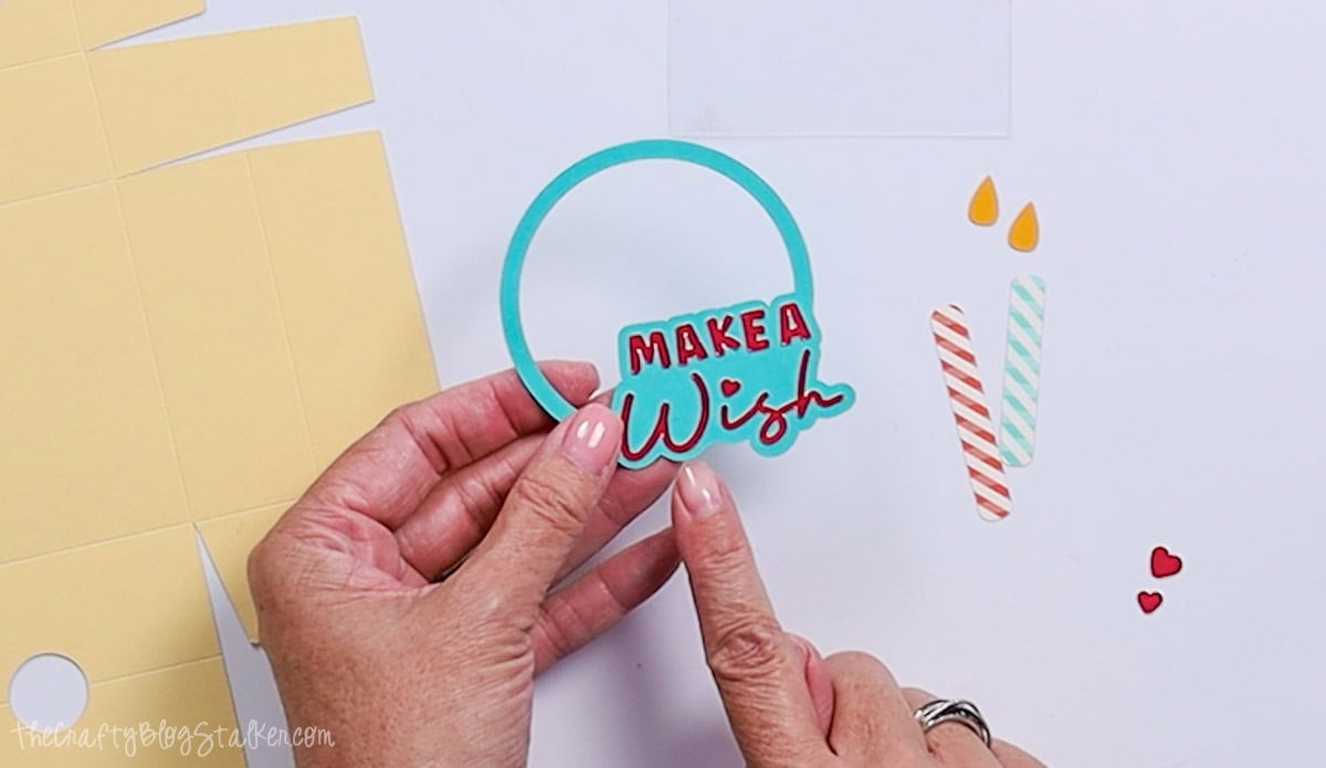 Hands hold a teal paper circle with a red “Make a Wish” design, ready to be assembled with candle and heart cutouts for a birthday craft project.