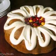 Pumpkin bundt cake with cream cheese frosting drizzled in a decorative pattern, topped with a center filled with red, yellow, and orange M&M candies on a dark red plate, with a stack of white plates and forks in the background.