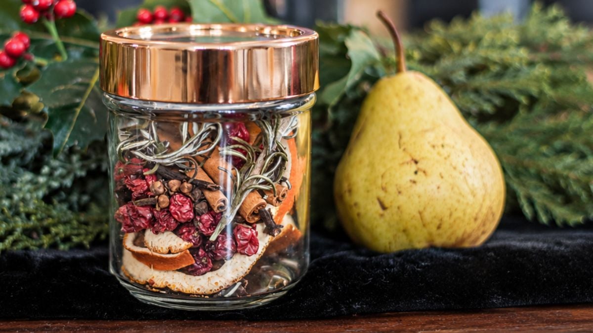 Christmas potpourri in a glass jar with dried oranges, cranberries, cinnamon sticks, cloves, and rosemary, displayed beside a fresh pear.