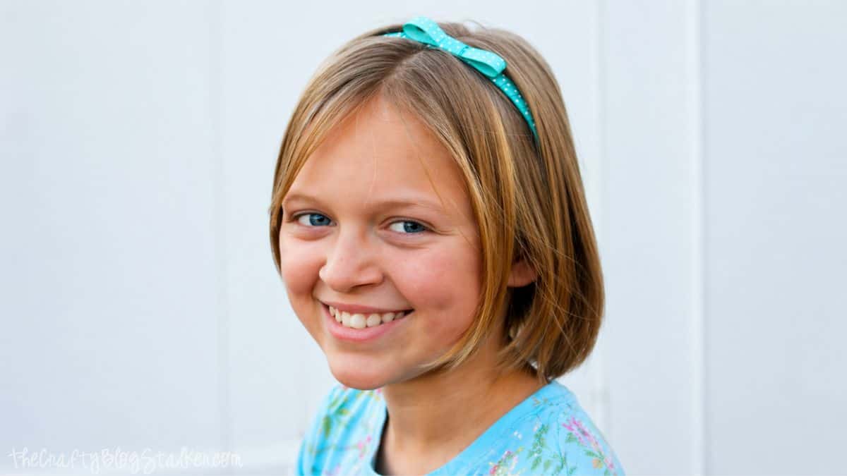 Smiling young girl with short hair wearing a teal bow headband and blue shirt, photographed against a light background.