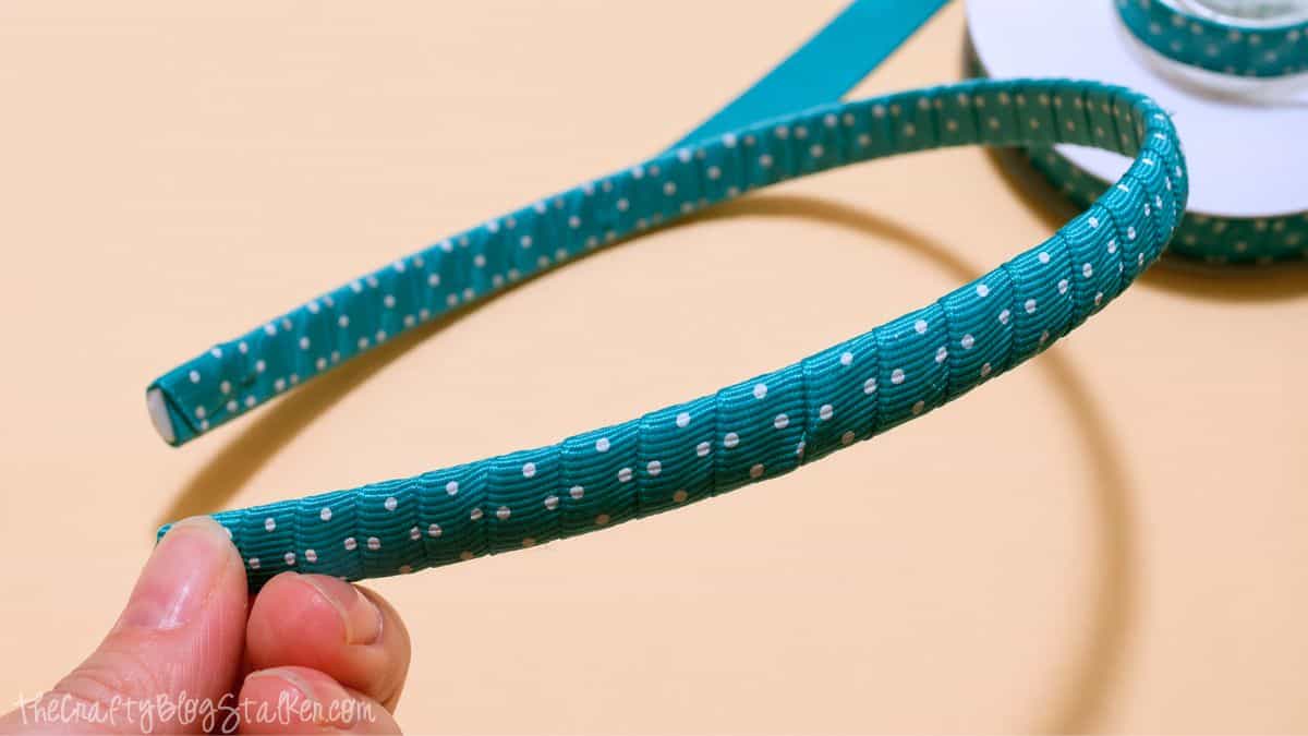 Hand holding a plastic headband wrapped in teal ribbon with white dots, with a ribbon spool in the background.