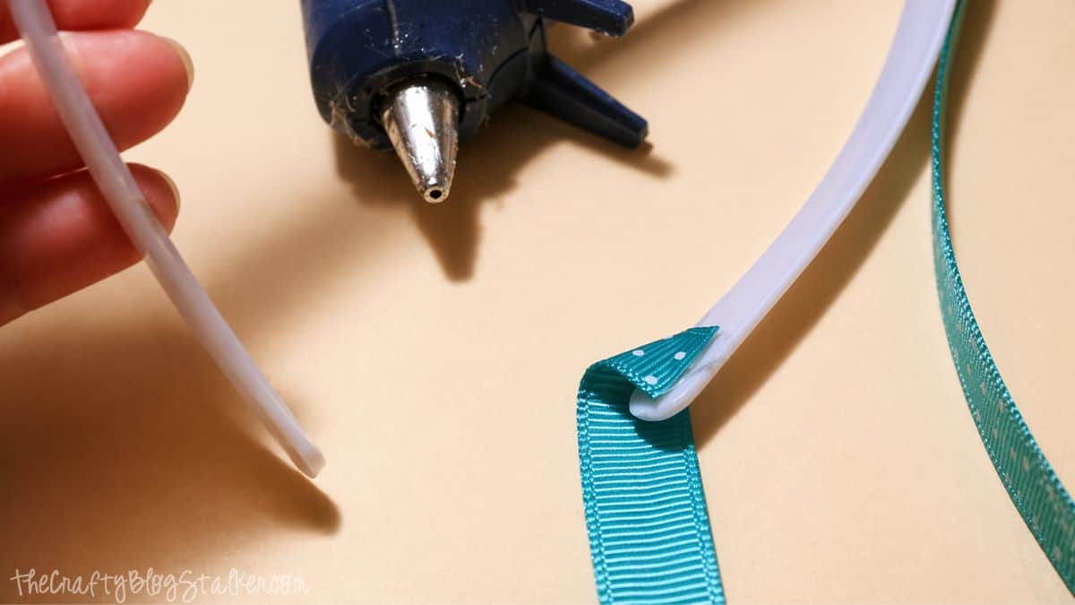 Hot glue gun applying ribbon to a plastic headband while a hand holds a glue stick during a DIY craft.