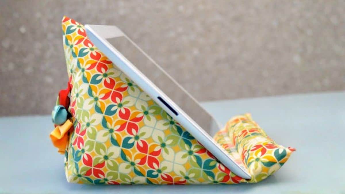 A handmade fabric iPad stand with a padded triangular shape, holding a tablet upright on a table for hands-free viewing or reading.