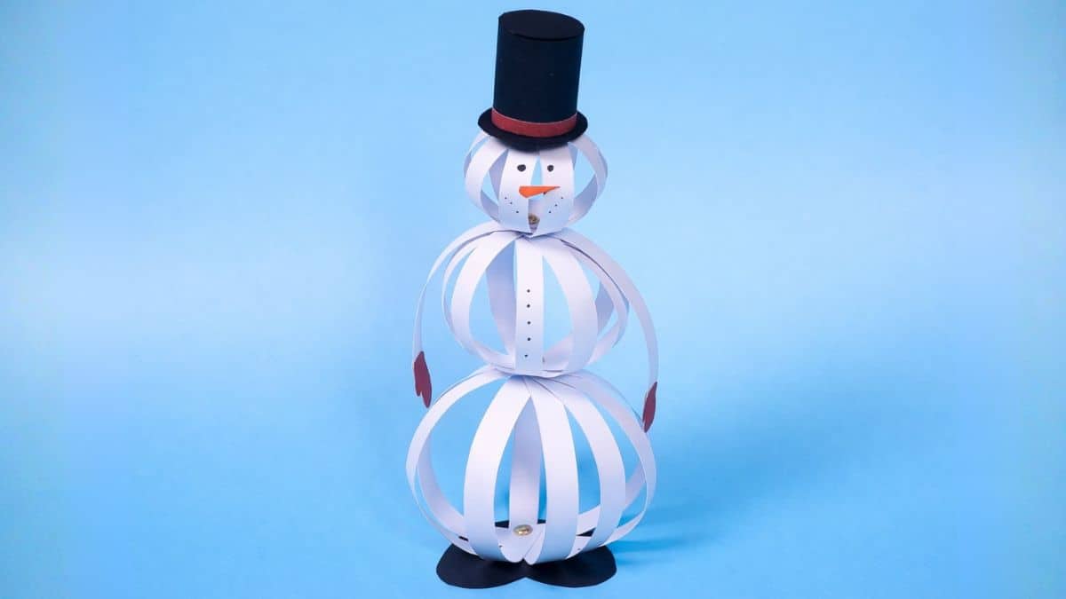Paper strip snowman craft standing upright, made from layered paper strips with a top hat, carrot nose, and mitten hands on a plain background.