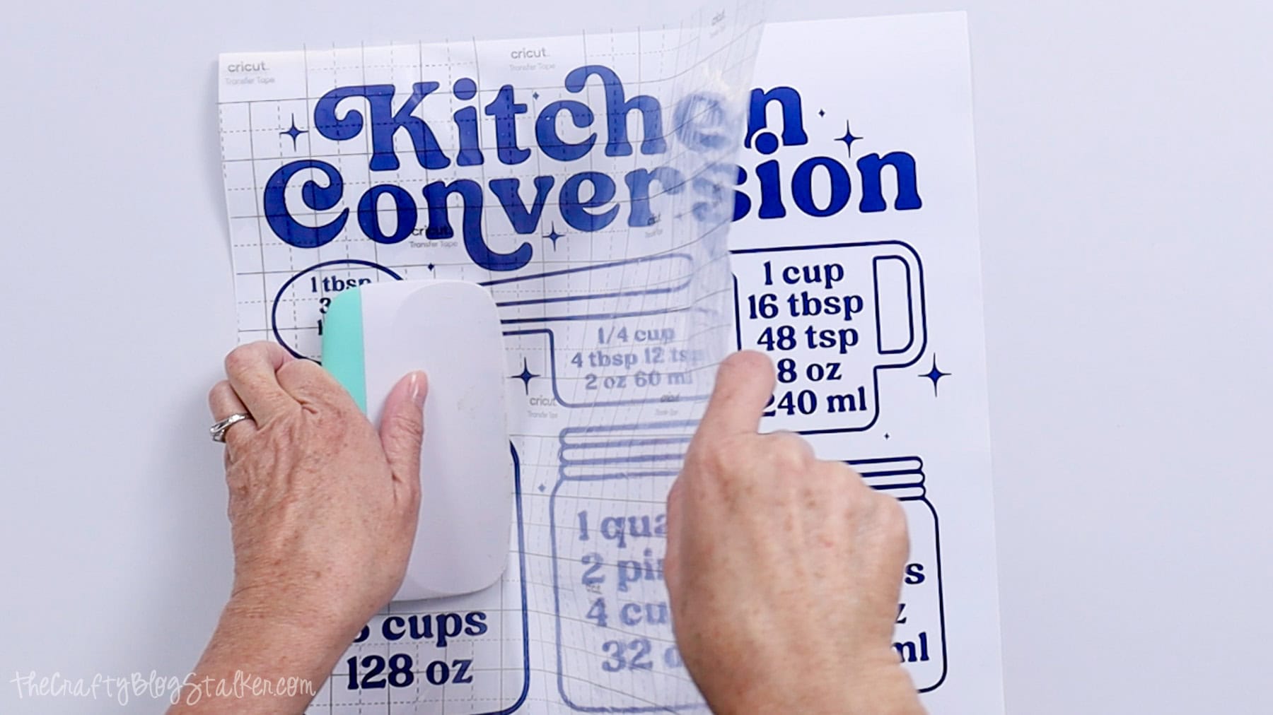 Hands using a scraper tool to apply transfer tape over a blue kitchen conversion chart vinyl decal.