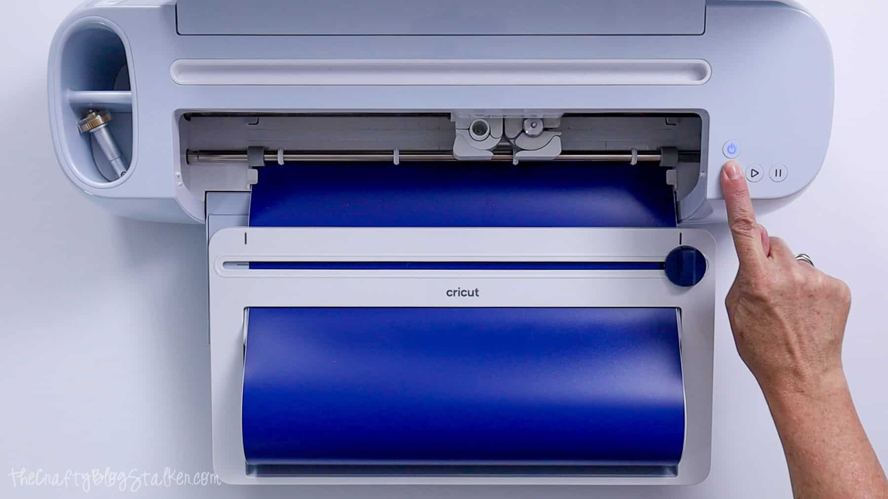 Hand pressing the start button on a Cricut machine as it cuts blue vinyl for a kitchen conversion chart decal.