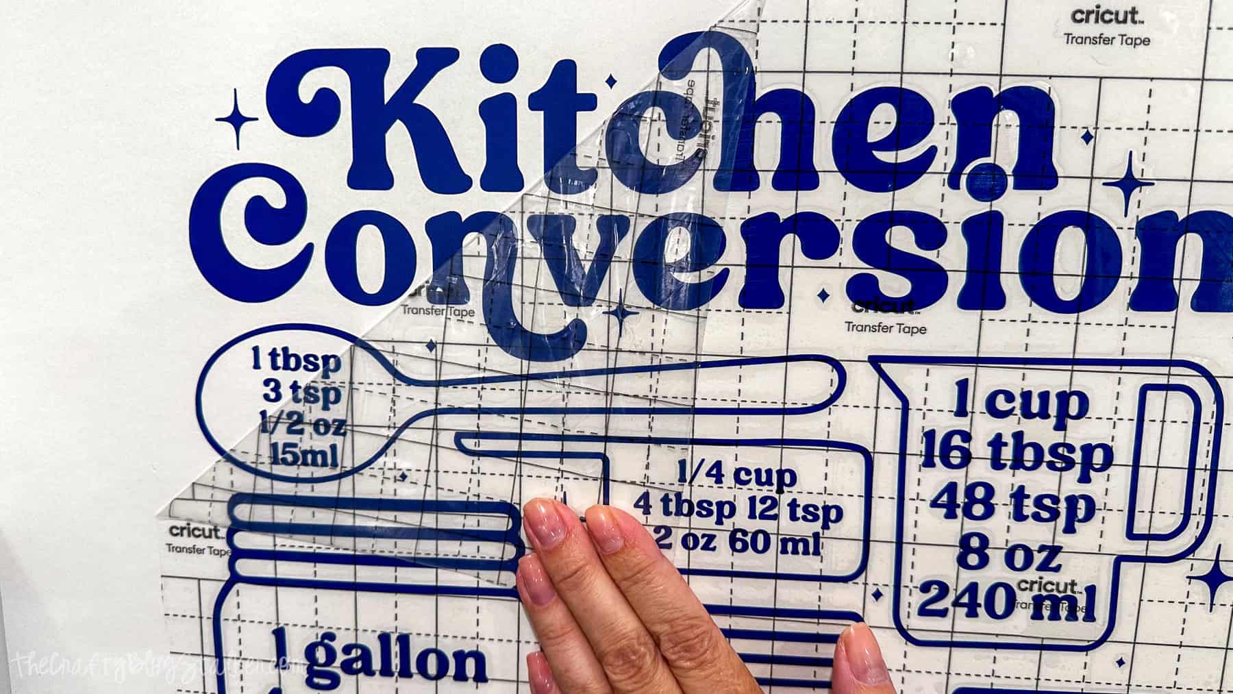 Hand peeling away transfer tape from a blue kitchen conversion chart vinyl decal after applying it to the wall.