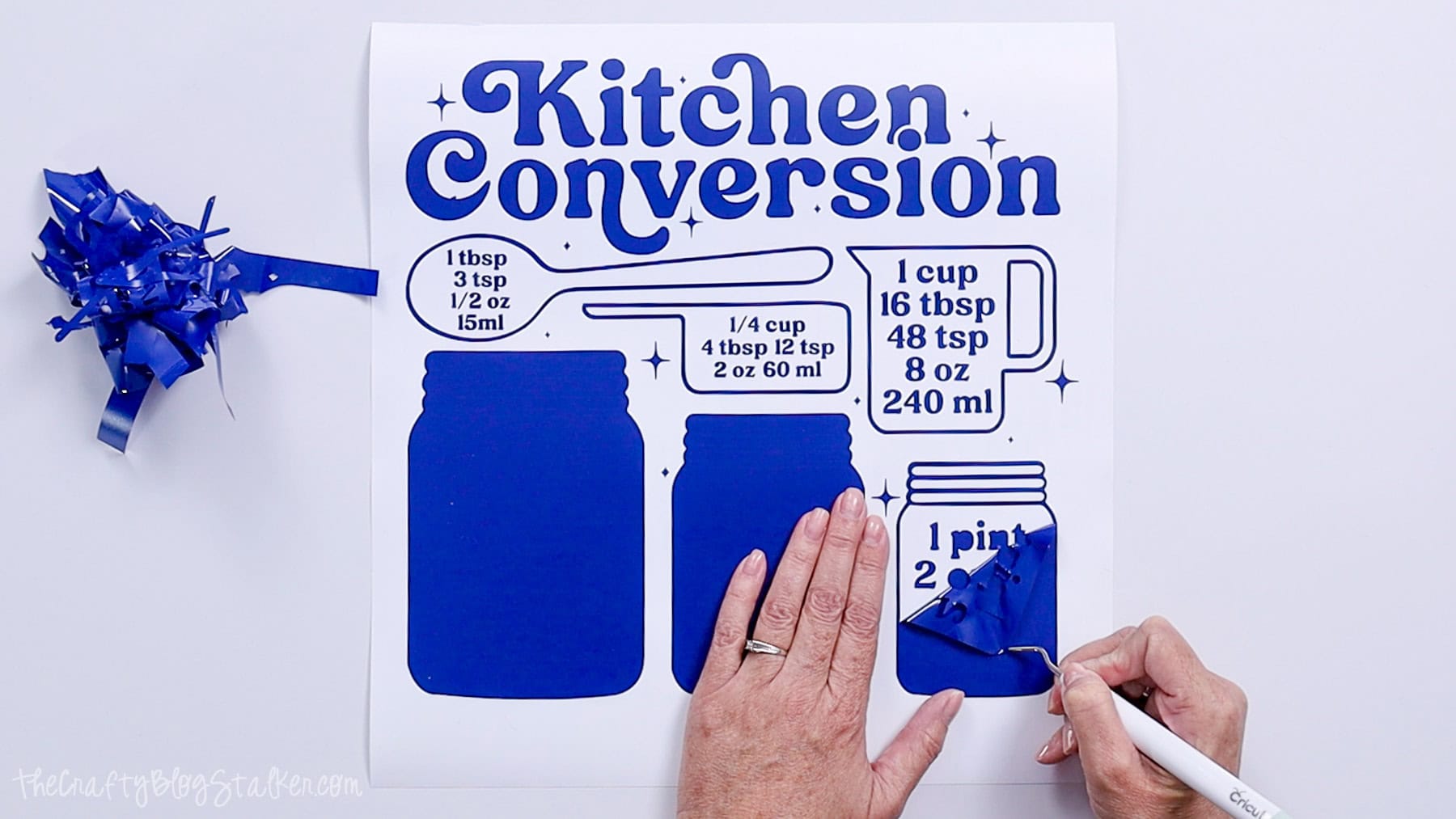 Hands using a Cricut weeding tool to remove excess vinyl from a blue kitchen conversion chart decal design.
