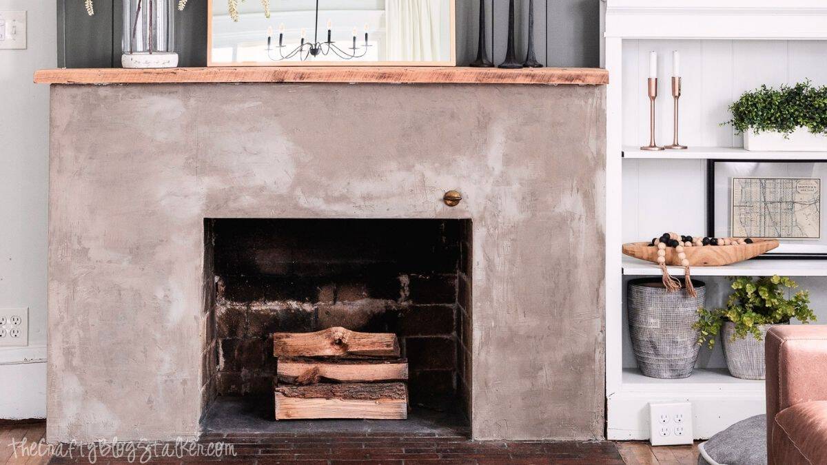 Concrete fireplace makeover with a smooth gray finish, natural wood mantel, stacked firewood, and styled built-in shelves on each side.