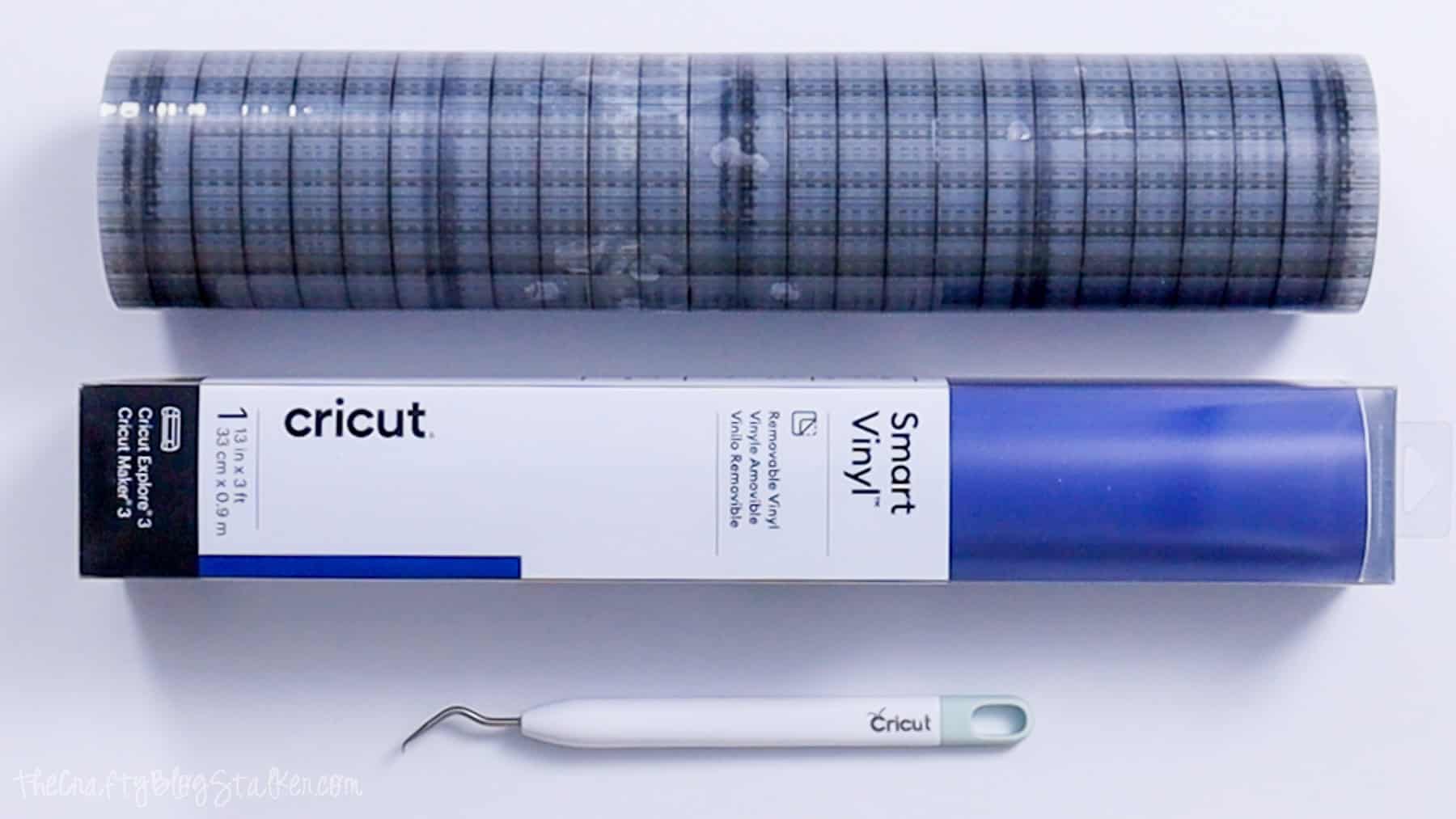 Cricut Smart Vinyl roll, transfer tape, and a weeding tool arranged on a white surface for a vinyl crafting project.