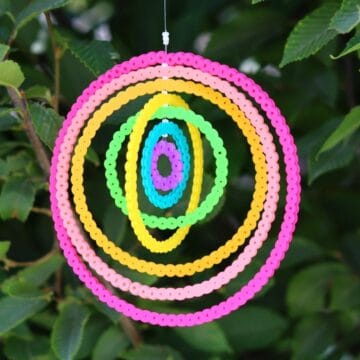 Colorful concentric circles made from Perler beads hanging outdoors in front of green leaves.