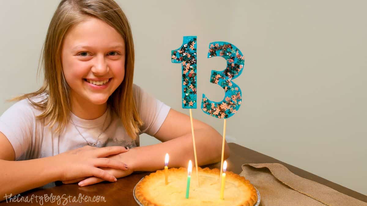 Smiling girl celebrates her 13th birthday with a pie topped with lit candles and a glittery blue “13” cake topper.