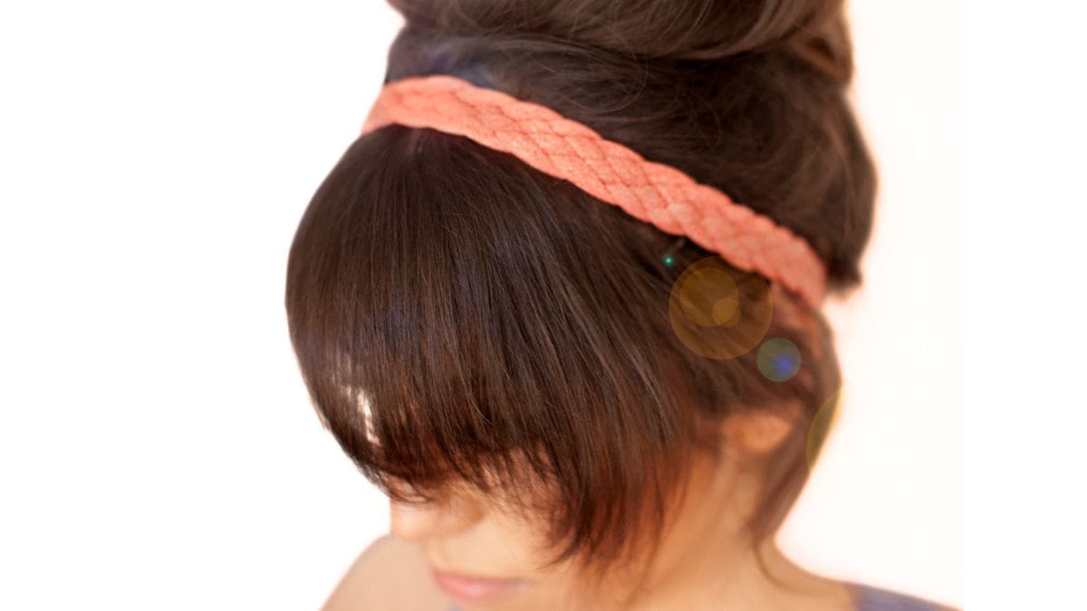 Woman with dark hair wearing a peach braided workout headband with bangs and hair styled in a messy bun.